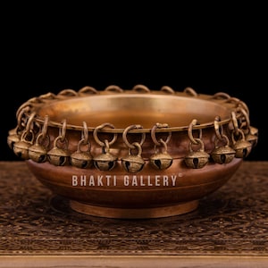 Op de afbeelding: Een kleine, ronde koperen kom met een decoratieve rand van kleine bellen. De kom staat op een gepatroneerd oppervlak. De tekst "BHAKTI GALLERY" is op de kom gedrukt.