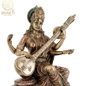 May include: A gold-toned statue of Saraswati, the Hindu goddess of knowledge, music, and art. She is depicted playing a sitar, a traditional Indian musical instrument. The statue is sitting on a rock base and has a detailed design.