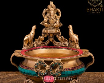 Puede incluir: Quemador de incienso de latón con un diseño detallado, con una pequeña estatua de Ganesha en la parte superior. El quemador está decorado con piedras preciosas rojas y verdes y tiene tres patas.