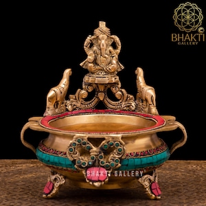 May include: Brass incense burner with an intricate design, featuring a small statue of Ganesha on top. The burner is decorated with red and green gemstones and has three legs.
