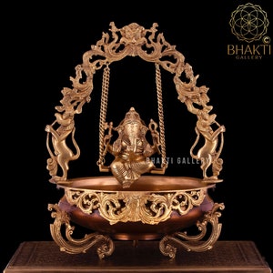 Może przedstawiać: Złota rzeźba z brązu przedstawiająca Ganesha, hinduskiego boga mądrości i dobrobytu, siedzącego na huśtawce. Huśtawka jest zawieszona na ozdobnym łuku, który jest podtrzymywany przez dwie figury lwów. Rzeźba stoi na okrągłym, miedzianym cokole z misternymi detalami.