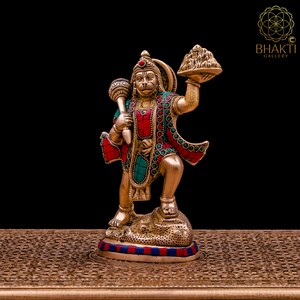 K&ouml;nnte beinhalten: Eine goldfarbene Statue der hinduistischen Gottheit Hanuman, die in der einen Hand einen Berg und in der anderen einen Kn&uuml;ppel h&auml;lt. Die Statue ist mit roten und t&uuml;rkisfarbenen Edelsteinen verziert.