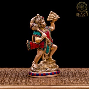 K&ouml;nnte beinhalten: Eine goldfarbene Statue der hinduistischen Gottheit Hanuman, die in ihrer rechten Hand einen Berg h&auml;lt. Die Statue ist mit roten und gr&uuml;nen Edelsteinen verziert und steht auf einem goldfarbenen Sockel.