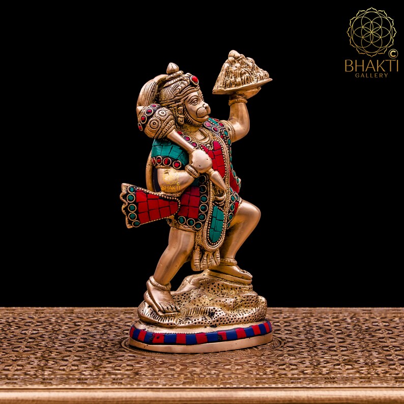 K&ouml;nnte beinhalten: Eine goldfarbene Statue der hinduistischen Gottheit Hanuman, die in ihrer rechten Hand einen Berg h&auml;lt. Die Statue ist mit roten und gr&uuml;nen Edelsteinen verziert und steht auf einem goldfarbenen Sockel.