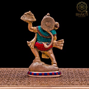 K&ouml;nnte beinhalten: Eine goldfarbene Statue von Hanuman, einer hinduistischen Gottheit, die einen Berg in seiner rechten Hand h&auml;lt. Die Statue ist mit t&uuml;rkisfarbenen und roten Edelsteinen verziert. Die Statue steht auf einem runden Sockel mit einem blauen und roten Rand.
