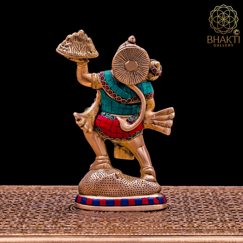 K&ouml;nnte beinhalten: Eine goldfarbene Statue von Hanuman, einer hinduistischen Gottheit, die einen Berg in seiner rechten Hand h&auml;lt. Die Statue ist mit t&uuml;rkisfarbenen und roten Edelsteinen verziert. Die Statue steht auf einem runden Sockel mit einem blauen und roten Rand.