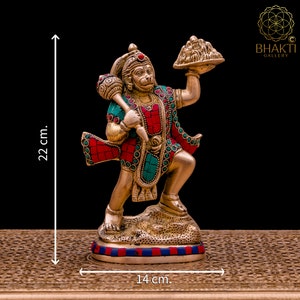 K&ouml;nnte beinhalten: Eine goldfarbene Statue der hinduistischen Gottheit Hanuman, die in der rechten Hand einen Berg und in der linken Hand eine Keule h&auml;lt. Die Statue ist mit roten und gr&uuml;nen Edelsteinen verziert und steht auf einem Sockel mit blauen und roten Streifen. Die Statue ist 22 cm hoch und 14 cm breit.