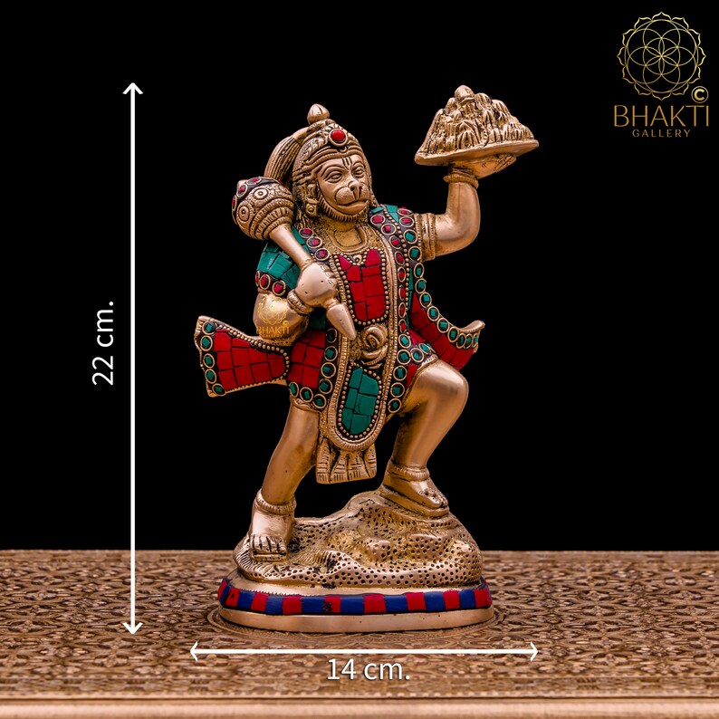 K&ouml;nnte beinhalten: Eine goldfarbene Statue der hinduistischen Gottheit Hanuman, die in der rechten Hand einen Berg und in der linken Hand eine Keule h&auml;lt. Die Statue ist mit roten und gr&uuml;nen Edelsteinen verziert und steht auf einem Sockel mit blauen und roten Streifen. Die Statue ist 22 cm hoch und 14 cm breit.
