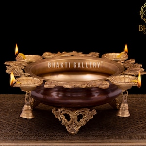 Może przedstawiać: Mosiężna diya z czterema knotami, każdy z płomieniem. Diya ma okrągłą miskę z ozdobną krawędzią i trzema nogami. Diya stoi na brązowej powierzchni z złotym wzorem.