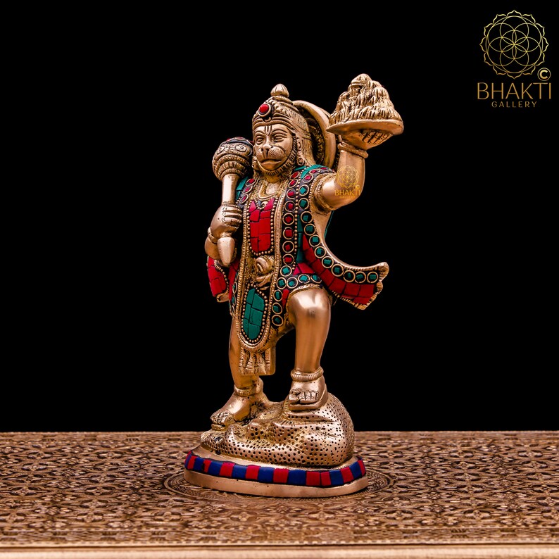K&ouml;nnte beinhalten: Eine goldfarbene Statue der hinduistischen Gottheit Hanuman, die in der einen Hand einen Kn&uuml;ppel und in der anderen eine Schale mit Fr&uuml;chten h&auml;lt. Die Statue ist mit roten und gr&uuml;nen Edelsteinen verziert und steht auf einem goldfarbenen Sockel.