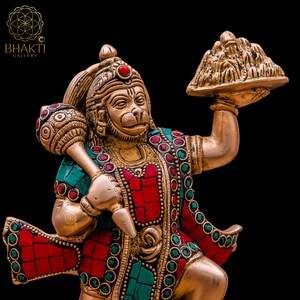 K&ouml;nnte beinhalten: Eine goldfarbene Statue der hinduistischen Gottheit Hanuman, die in der einen Hand eine Keule und in der anderen eine Schale mit Fr&uuml;chten h&auml;lt. Die Statue ist mit roten und gr&uuml;nen Edelsteinen verziert.