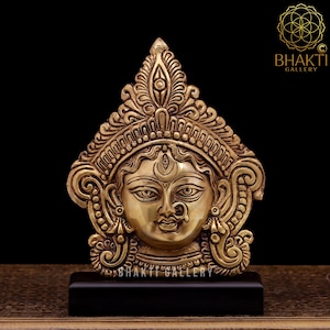 Puede incluir: Una escultura de metal dorado de una diosa hindú con intrincados detalles. La escultura está sobre una base de madera negra. La escultura es una representación de la diosa hindú Durga.