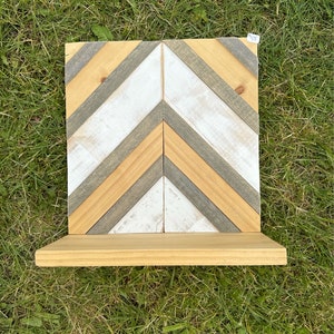 Op de afbeelding: Een houten plank met een visgraatpatroon. De plank is gemaakt van verschillende houttinten, waaronder wit, grijs en natuurlijk hout. De plank heeft een kleine, lichtgekleurde plank die zich vanaf de onderkant uitstrekt. De plank ligt op groen gras.