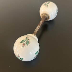 May include: A vintage white ceramic door knob with a floral design featuring red buds and green leaves. The knob is connected by a rusty metal rod. The overall design is a classic, antique style.