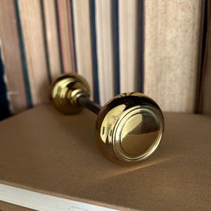 May include: A polished brass door knob featuring a smooth, rounded face and a stepped circular base. The knob is connected to a central metal spindle, showcasing a reflective metallic finish that catches the light across its curved surface.