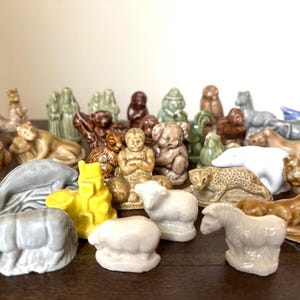 Könnte beinhalten: Eine Sammlung kleiner Keramik-Tierfiguren in verschiedenen Farben, darunter Braun, Grau, Gelb und Weiß. Die Figuren stellen Tiere wie Löwen, Delfine und Bären dar. Die Sammlung ist auf einer dunklen Holzoberfläche angeordnet.