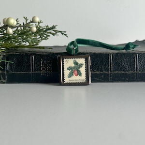 May include: A vintage Christmas ornament featuring a 5-cent United States postage stamp with a pine bough and red berry design. The stamp is set in a rectangular metal frame with a green velvet ribbon for hanging.
