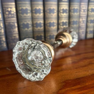 May include: A clear, faceted glass door knob with a fluted, starburst-like pattern on the face. The knob features a circular brass-toned metal base and a central metal spindle connecting it to a matching glass knob in the background.