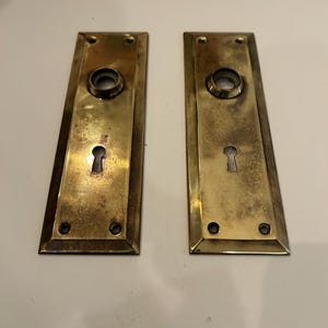 May include: Two antique brass door plates. Each plate has a keyhole, a circular opening, and screw holes. The brass has a warm, aged patina, suggesting vintage or historical use. The plates are rectangular with a slightly tapered design.