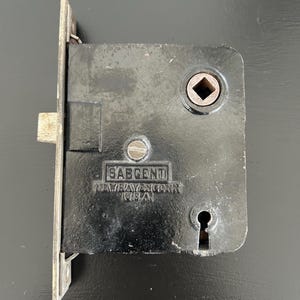 May include: A black metal door lock with a keyhole and a square-shaped key slot. The lock is labeled "SARGENT NEW HAVEN, CONN. USA."