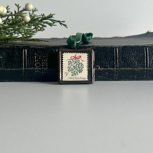 May include: A small, rectangular, framed vintage postage stamp featuring a green mistletoe design with a red bow. The stamp is set in a bronze-colored frame with a green velvet ribbon. The text "United States Postage" is visible.
