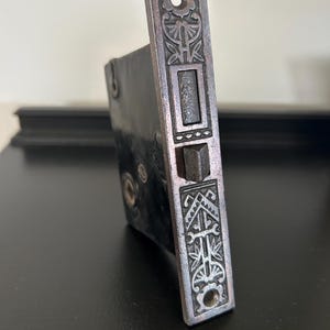 May include: An antique, rectangular door lock with ornate floral and geometric designs. The lock is made of dark metal with a slightly iridescent finish. The design includes a rectangular latch and decorative screw holes, showcasing a vintage aesthetic.