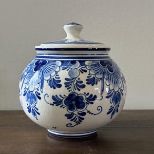 May include: A white ceramic jar with a lid, adorned with blue floral patterns. The jar has a round shape and sits on a wooden surface. The lid has a small knob for easy handling. The design is reminiscent of Delftware.