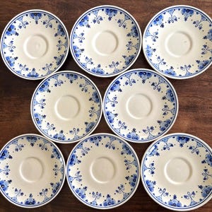 May include: A collection of eight off-white ceramic saucer plates with a blue floral design around the rim. The plates are arranged on a dark wooden surface. Each saucer has a central recessed area.