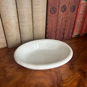May include: An oval-shaped, off-white ceramic dish with a smooth, glossy finish and a subtle crackle glaze texture. The small, shallow bowl features a rounded rim and a simple, clean design, presented as a standalone piece for various uses.