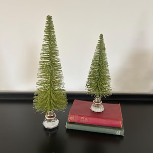 May include: Two green bottle brush Christmas trees with clear glass bases and gold accents. One tree is taller than the other. The trees are displayed on a black surface with two antique books.