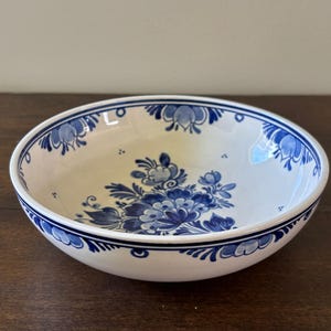 May include: A white ceramic bowl with a blue floral design. The bowl has a wide rim with a blue border and a central floral motif. The design features stylized flowers and leaves in shades of blue.