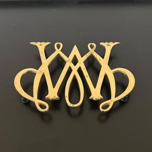 Może przedstawiać: Złota metalowa broszka z przeplatanym wzorem liter "W" i "M". Broszka ma dekoracyjny, ozdobny styl z polerowanym wykończeniem. Broszka jest umieszczona na czarnym tle.