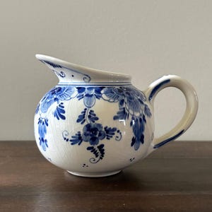 May include: A small, round, white ceramic pitcher with a curved spout and handle. The pitcher is decorated with blue floral patterns and accents. The design includes flowers, leaves, and other botanical elements. The pitcher sits on a dark wooden surface.