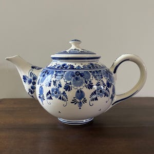 May include: A white ceramic teapot with a blue floral pattern. The teapot has a rounded body, a curved spout, and a loop handle. The lid is topped with a small knob. The design is a classic style, suitable for serving tea.