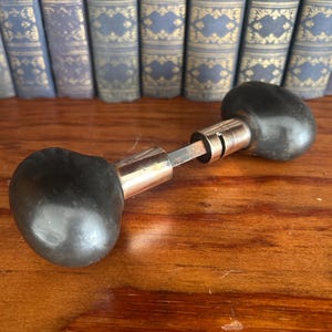 May include: A door knob set featuring two smooth, rounded black knobs connected by a central metallic spindle. The hardware includes a polished silver-toned cylindrical neck piece attached to each knob, providing a functional mechanism for door operation.