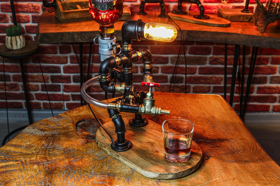 May include: A black metal pipe lamp with a clear glass bulb and a clear glass shot glass. The lamp is shaped like a person and has a red valve on the side. The lamp is sitting on a wooden table with a brown wooden base.