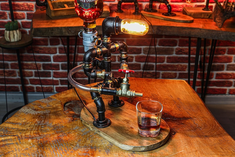 May include: A steampunk-style liquor dispenser made from black pipes and fittings, with a clear glass bottle, a red valve, and a clear glass shot glass. The dispenser has a light bulb with a warm glow.