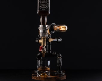 Puede incluir: Un dispensador de licor de metal negro con una botella de vidrio transparente, una válvula de latón y una bombilla. El dispensador está vertiendo licor en un vaso transparente.