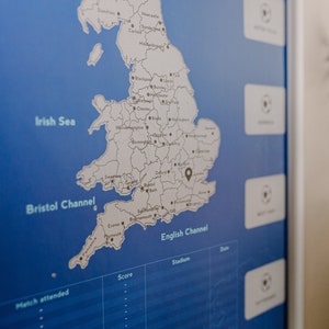 Peut inclure: Une carte bleue et blanche de l'Angleterre avec une &eacute;pingle noire marquant un emplacement dans le sud-est. La carte est sur un fond bleu avec une bordure blanche. La carte est &eacute;tiquet&eacute;e avec les noms des villes et des villages. La carte fait partie d'un tableau plus grand avec des colonnes intitul&eacute;es "Score", "Stade" et "Date".