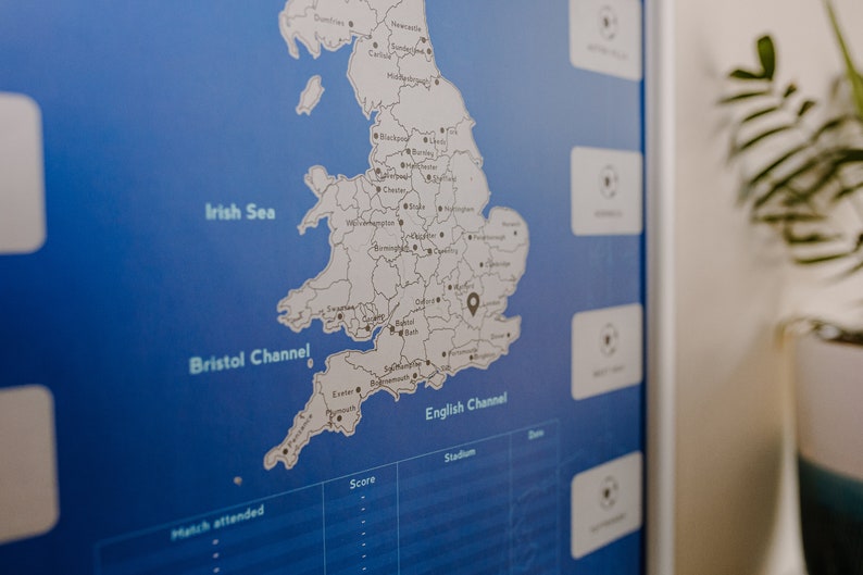 Peut inclure: Une carte bleue et blanche de l'Angleterre avec une &eacute;pingle noire marquant un emplacement dans le sud-est. La carte est sur un fond bleu avec une bordure blanche. La carte est &eacute;tiquet&eacute;e avec les noms des villes et des villages. La carte fait partie d'un tableau plus grand avec des colonnes intitul&eacute;es "Score", "Stade" et "Date".