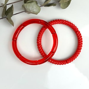 Könnte beinhalten: Zwei leuchtend rote Armreifen. Einer hat ein glattes, segmentiertes Design, der andere eine gedrehte, seilartige Textur. Die Armreifen werden auf einer weißen Oberfläche mit einem Zweig Grünzeug präsentiert. Schmuck.