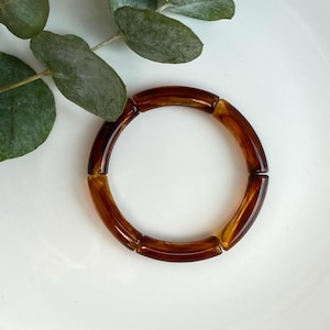 Könnte beinhalten: Ein braunes, segmentiertes Armband mit glänzendem Finish. Das Armband besteht aus einem durchscheinenden Material mit warmer, bernsteinfarbener Farbe. Das Armband ist kreisförmig und liegt auf einer weißen Oberfläche, mit grünen Blättern im Hintergrund.