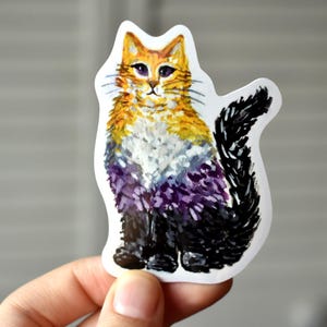 May include: A sticker featuring a watercolor illustration of a cat. The cat has a fluffy orange and yellow head, a white chest, and a purple and black body. The cat is sitting upright with its tail curled.