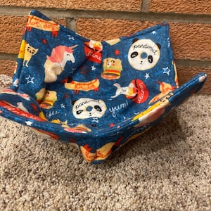 May include: A blue fabric bowl with a variety of colorful cartoon images, including a hot dog, a unicorn, a panda, and a hamburger. The bowl is lined with a white fabric and has a star pattern.