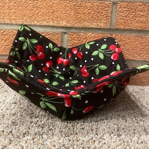 May include: A black fabric bowl with a cherry and white polka dot pattern. The bowl is folded in half and is sitting on a brown carpet.