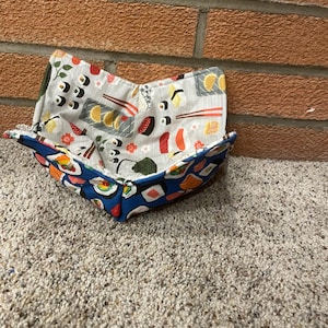 May include: A handmade fabric bowl with a sushi pattern. The bowl has a blue exterior with sushi illustrations and a white interior with sushi and chopstick designs. The bowl is sitting on a carpeted surface.