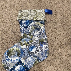 May include: A blue and green Christmas stocking with a white background and a blue ribbon. The stocking is decorated with a variety of patterns, including snowflakes, trees, and ornaments.
