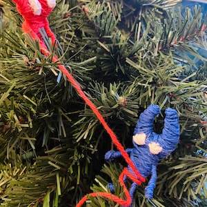 May include: Two yarn figures, one red and one blue, are climbing a green artificial Christmas tree. The red figure is at the top of the tree, while the blue figure is climbing up the tree.
