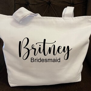 May include: A white tote bag with two handles. The name "Britney" is printed in a cursive font above the word "Bridesmaid" in a sans-serif font. The bag is made of a sturdy fabric.