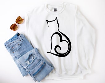 Peut inclure: Sweat-shirt blanc avec un motif de chat noir. Le corps du chat est formé par un cœur. Un jean bleu clair et des lunettes de soleil marron sont à côté du sweat-shirt.
